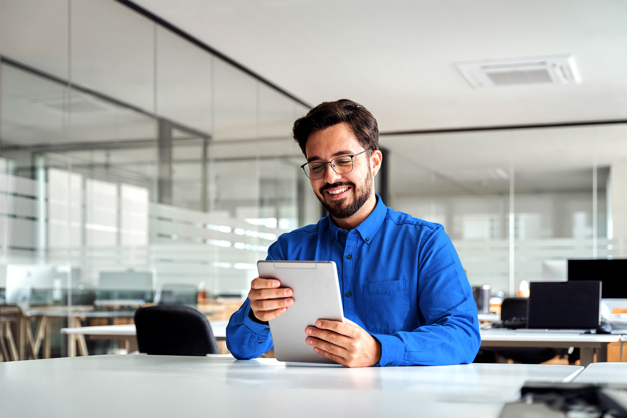 Man in blue shirt smiling while using a tablet in a modern office