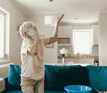 Senior homeowner on the phone reporting a ceiling water leak in a modern living room, showing urgenc…