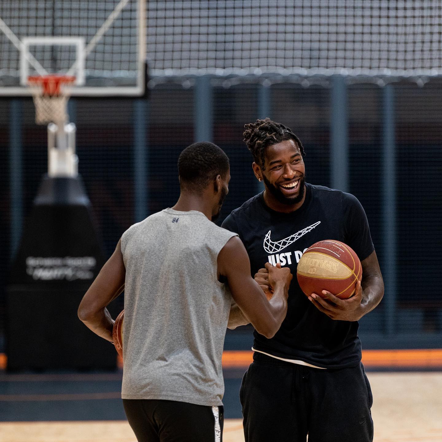 Deux hommes tenant un ballon de basket se saluent avec un sourire sur un terrain de basket en intérieur.