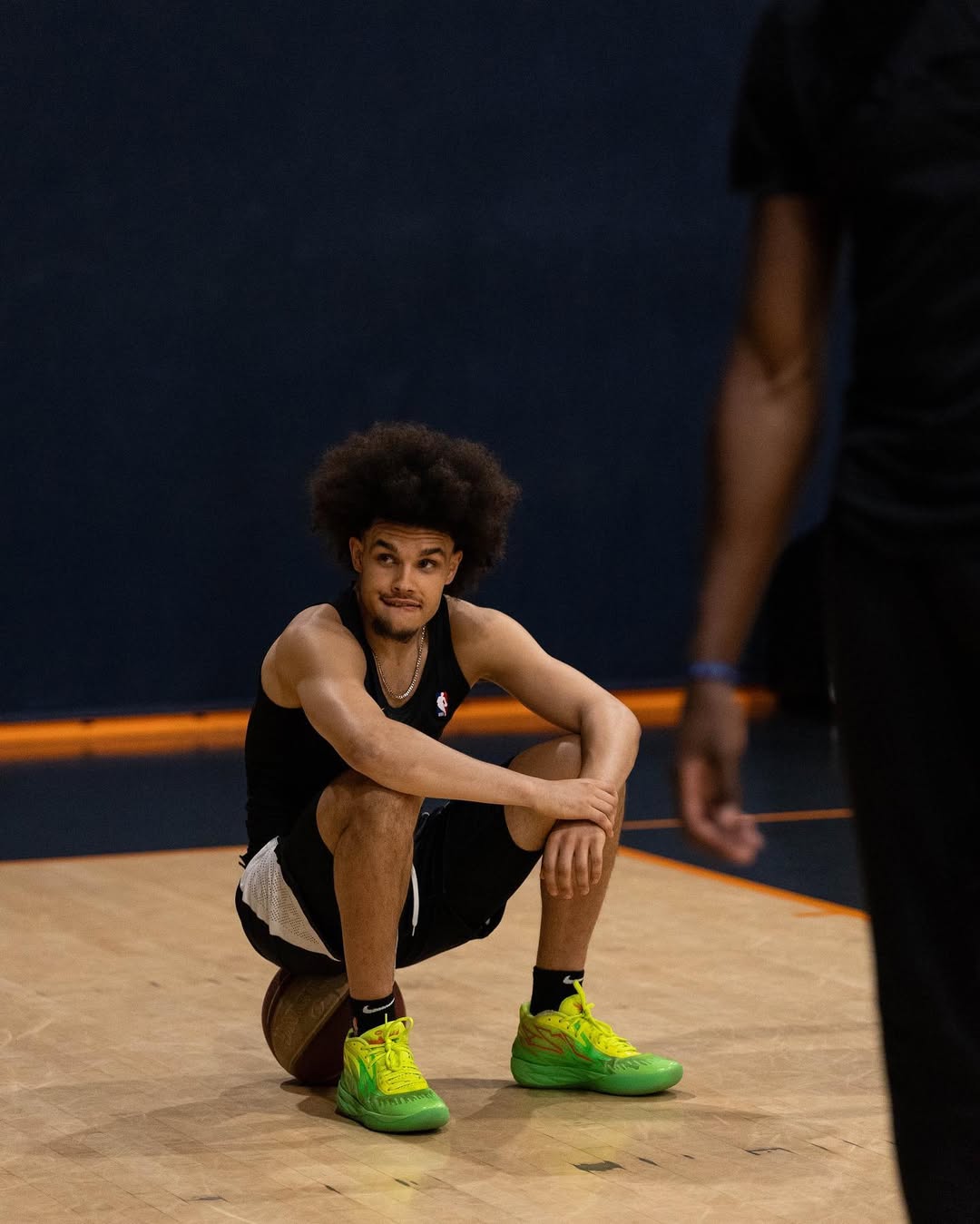 Jeune homme assis sur un ballon de basket sur un terrain intérieur, portant un débardeur noir, un short noir et blanc, et des baskets vertes vives.