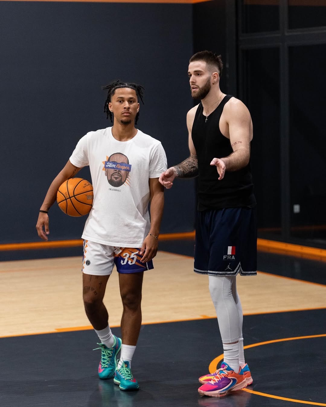 Deux joueurs de basketball en tenue sportive sur un terrain intérieur, l'un tenant un ballon, l'autre parlant.