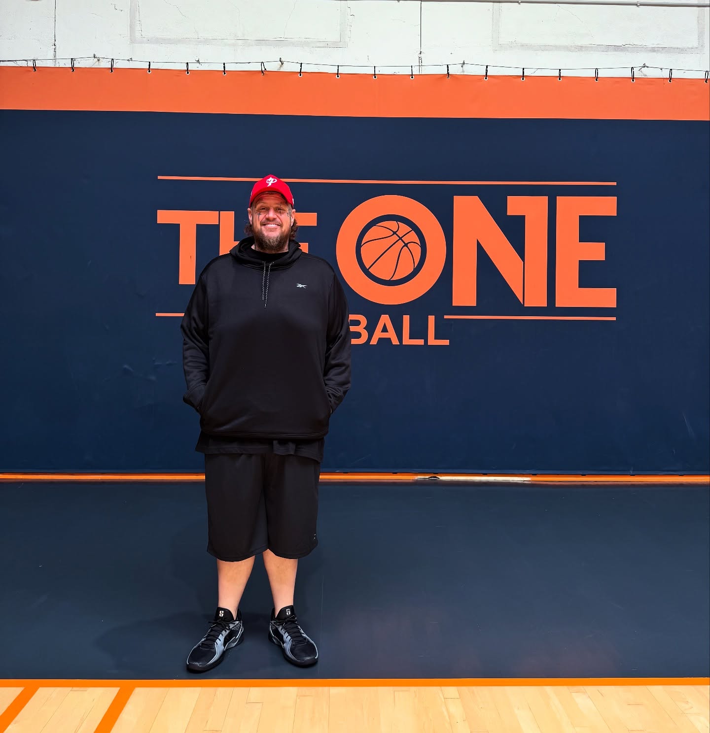 Un homme souriant portant un sweat à capuche noir, un short noir et une casquette rouge, debout sur un sol de gymnase devant un mur bleu avec le logo orange "THE ONE BASKETBALL".