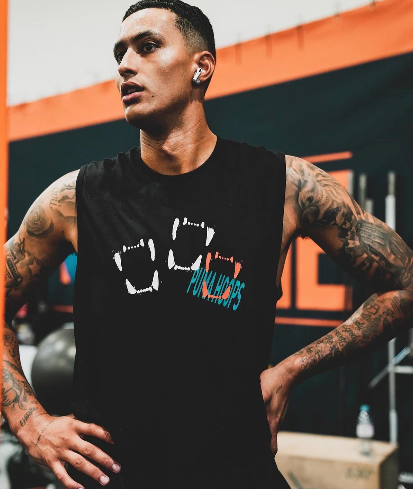 Homme tatoué portant un débardeur noir avec logo 'Puma Hoops' dans une salle de sport.