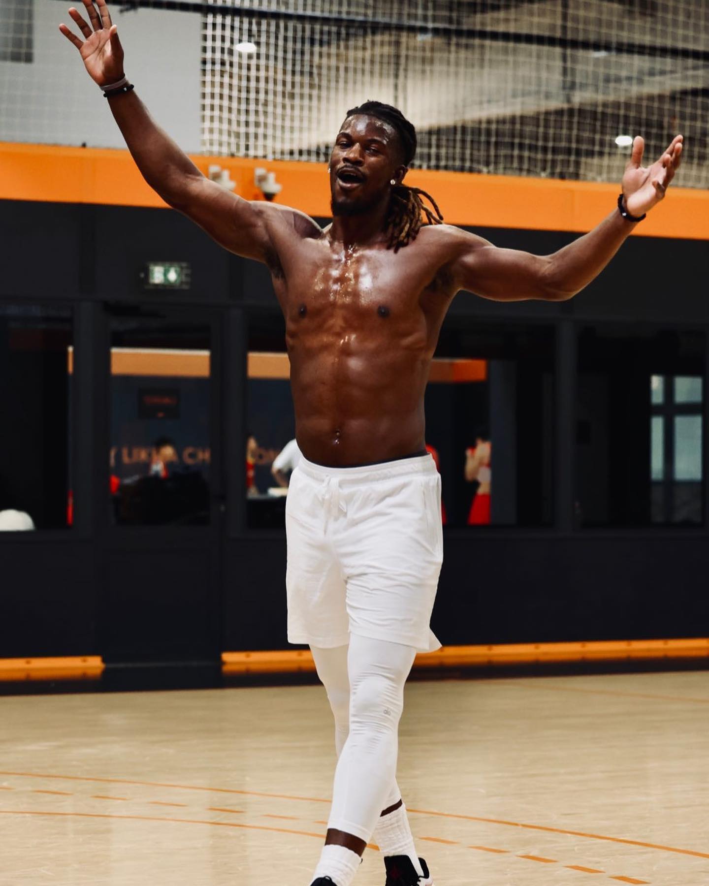 Homme musclé torse nu portant un short blanc et un legging blanc, bras écartés, sur un terrain de sport intérieur.