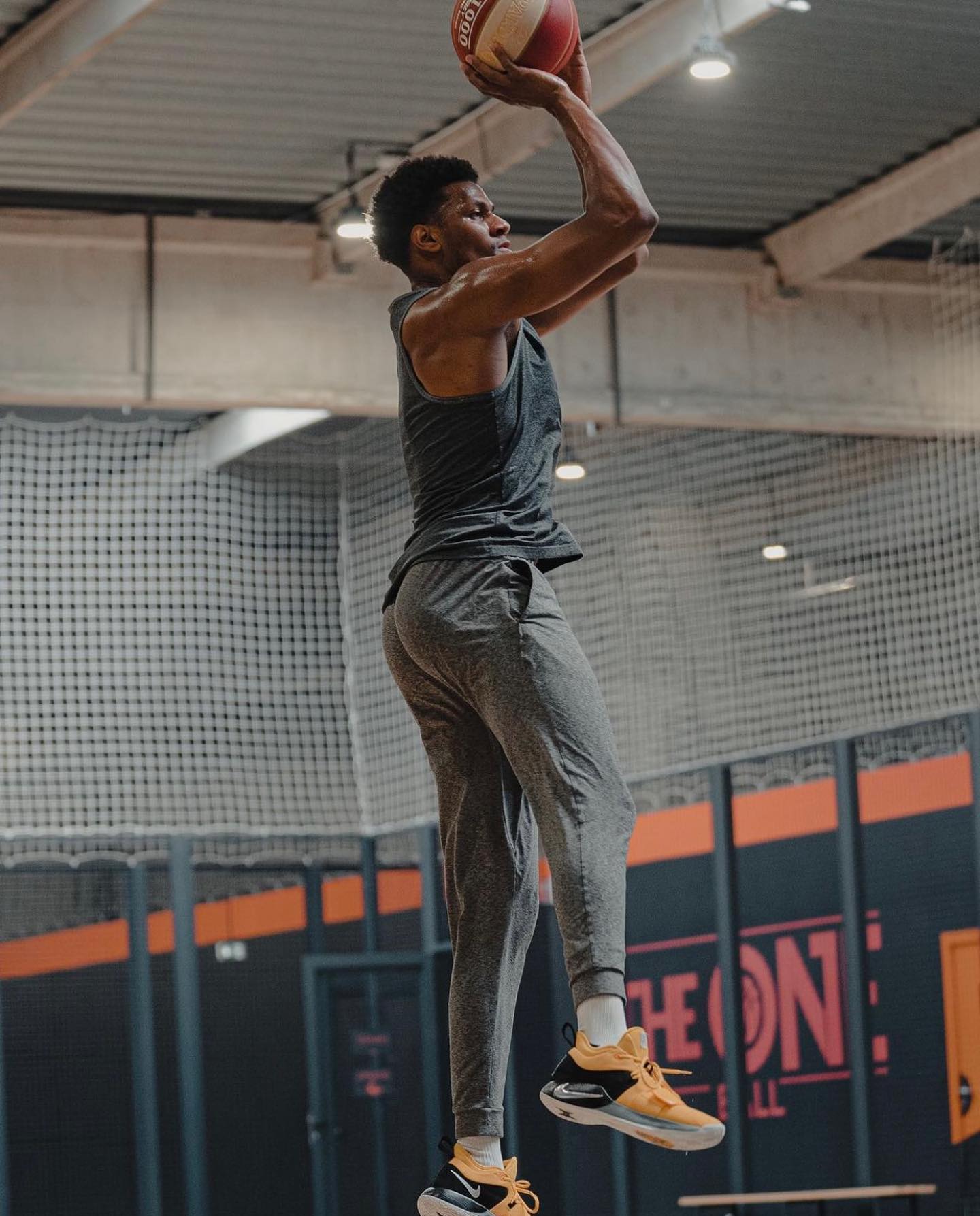 Un joueur de basketball en tenue sportive grise effectuant un saut pour tirer le ballon dans un gymnase.