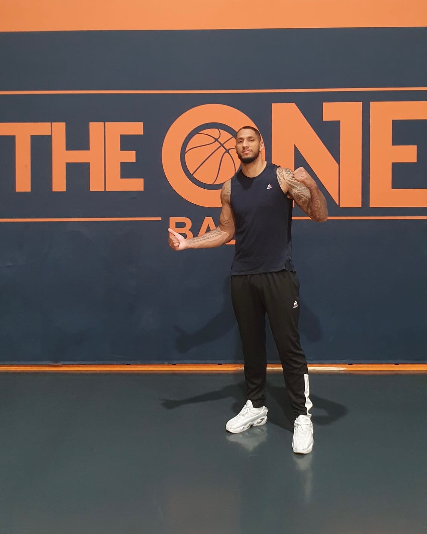 Homme debout devant un mur avec le logo 'THE ONE' en orange, portant un débardeur noir et un pantalon de sport.