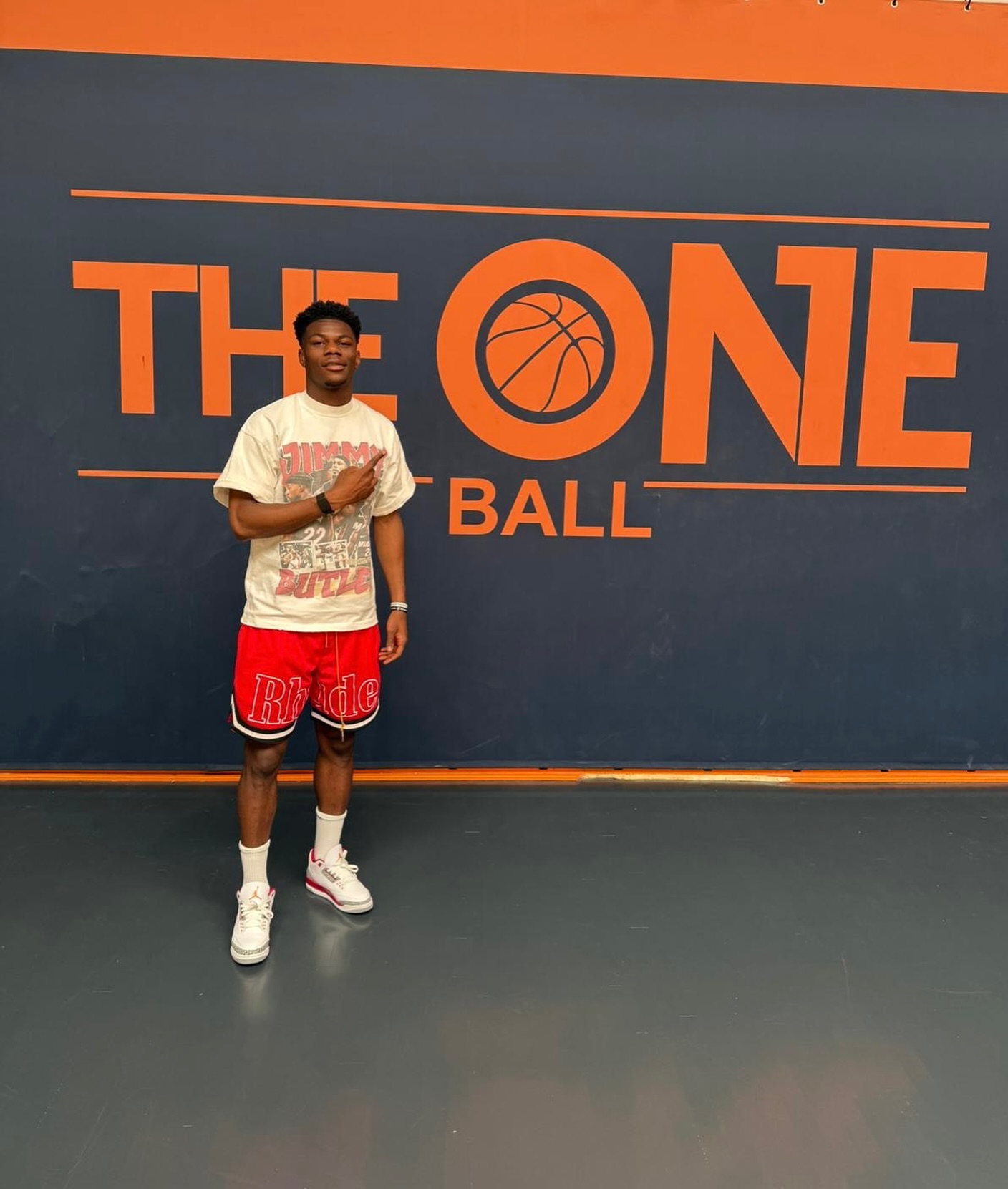 Un homme debout sur un terrain de basket indoor, pointant vers un mur avec le texte orange "THE ONE BALL" et un logo de ballon de basket.