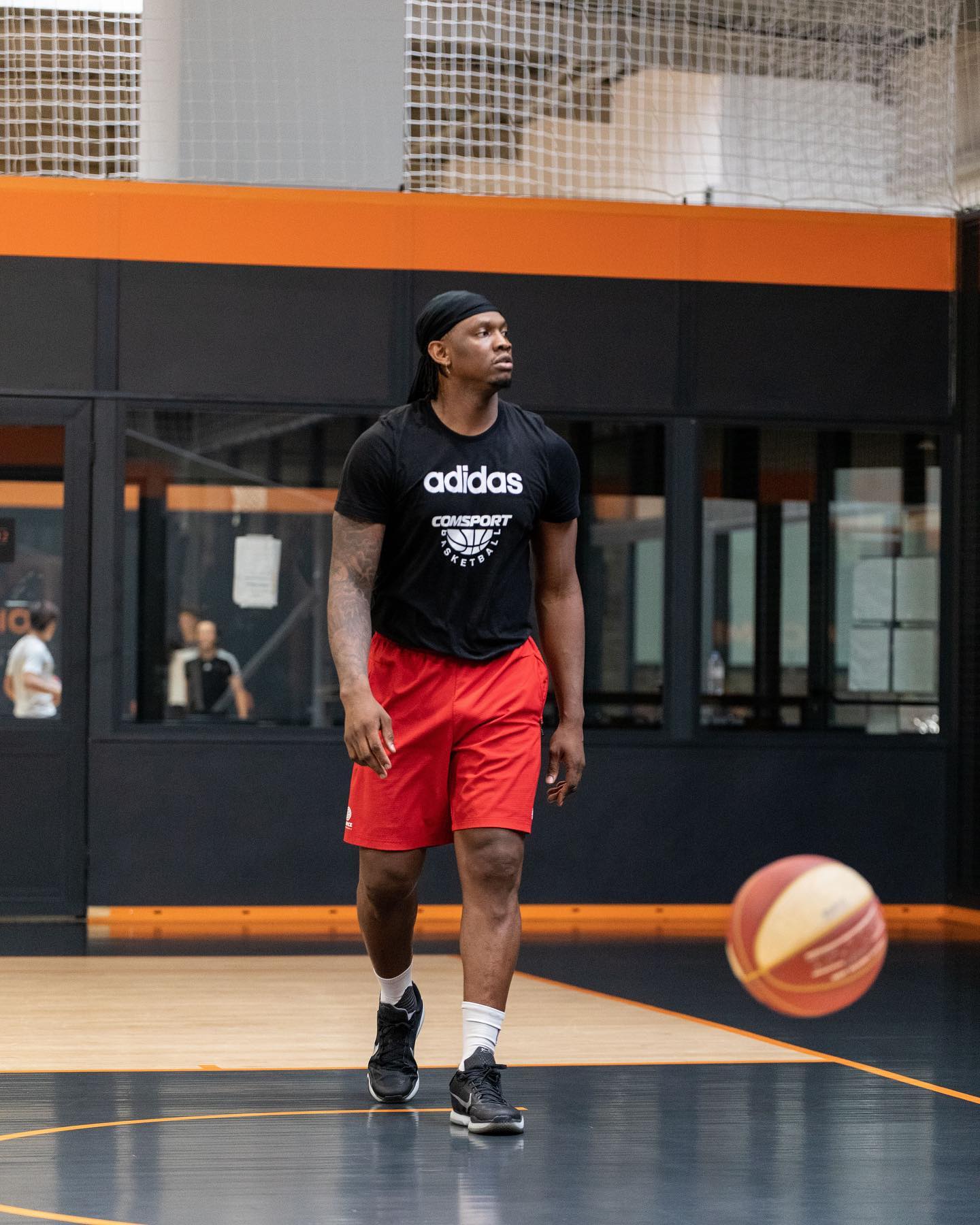 Un joueur de basket marche sur un terrain intérieur, portant un t-shirt noir Adidas, un short rouge et des baskets noires, avec un ballon de basket flou au premier plan.
