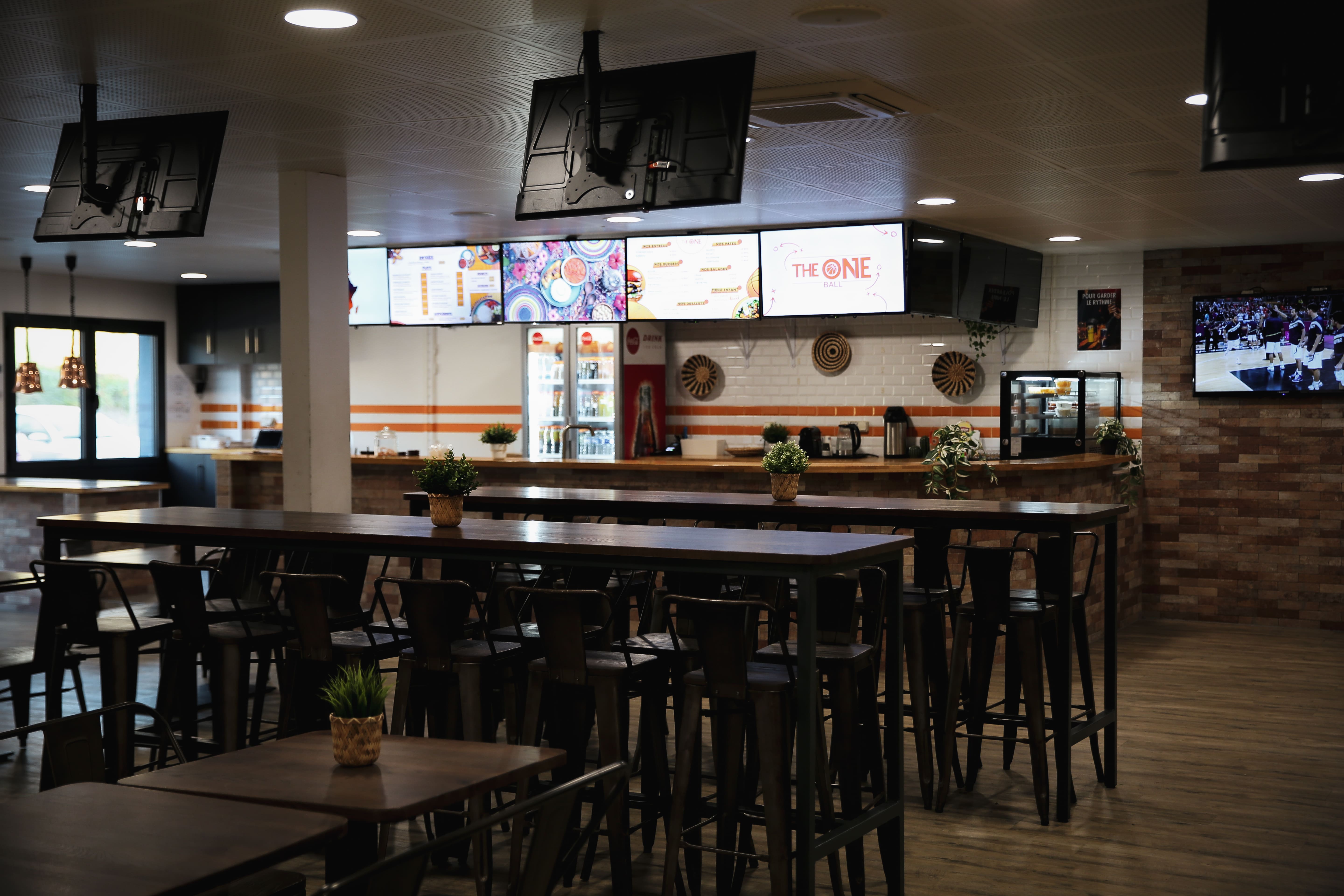Intérieur d'un restaurant moderne avec tables hautes, chaises noires, plantes décoratives, et plusieurs écrans numériques affichant un menu et une publicité.