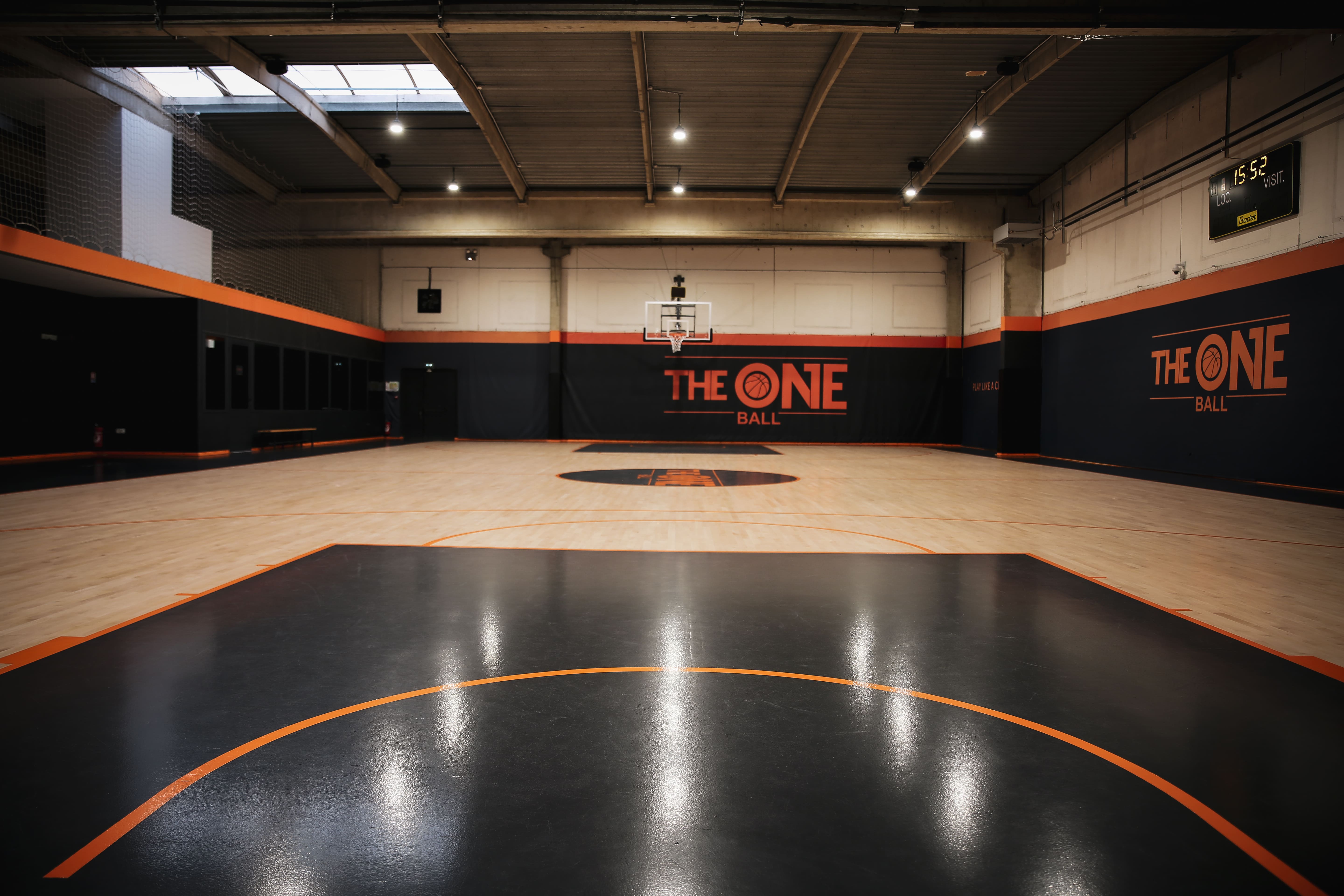 Terrain de basket-ball intérieur avec un sol clair et zone clé noire, murs ornés du logo orange 'The One Ball'.