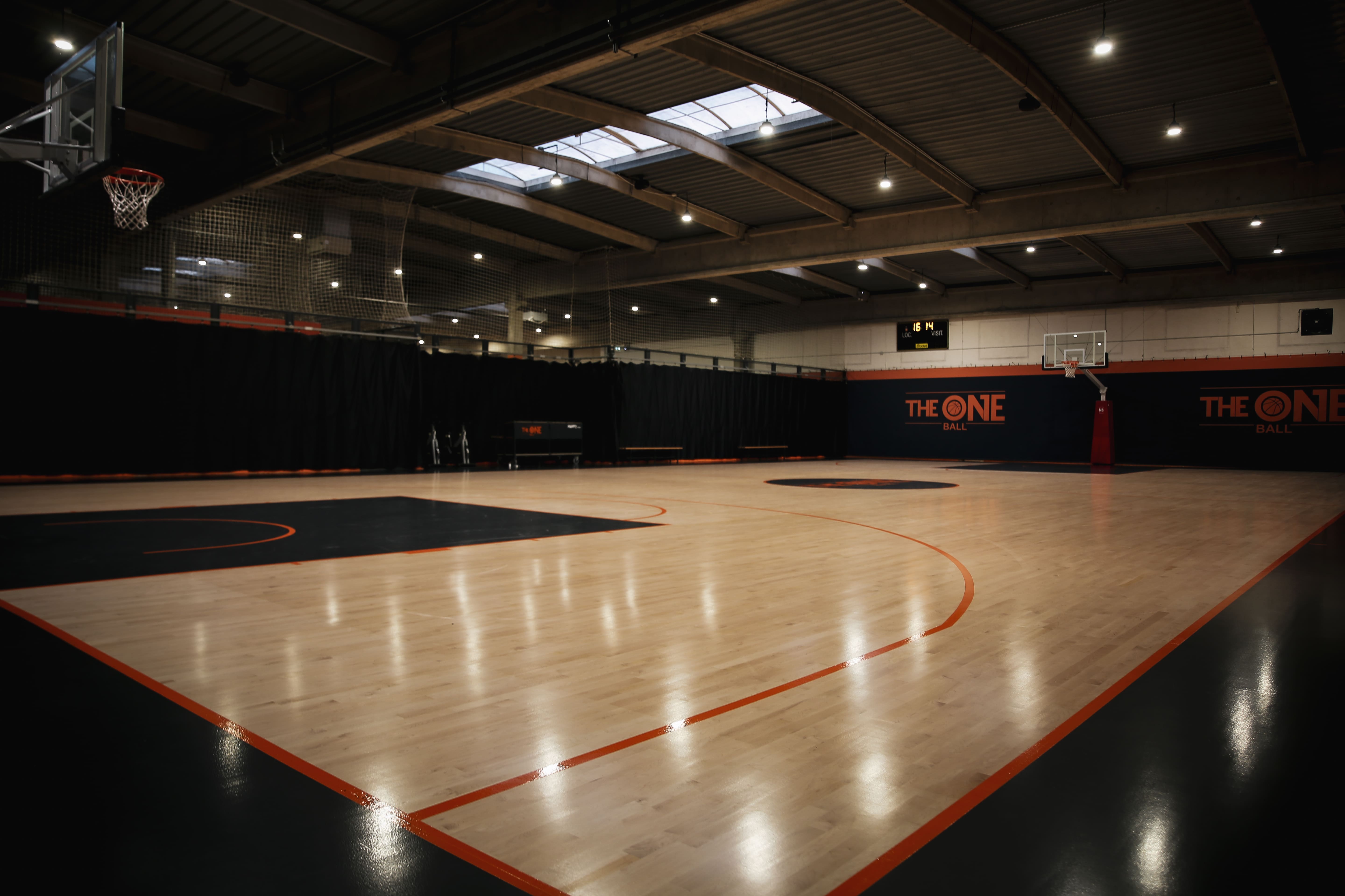 Terrain de basket-ball intérieur vide avec filets de protection et panneaux "THE ONE BALL" sur les murs.