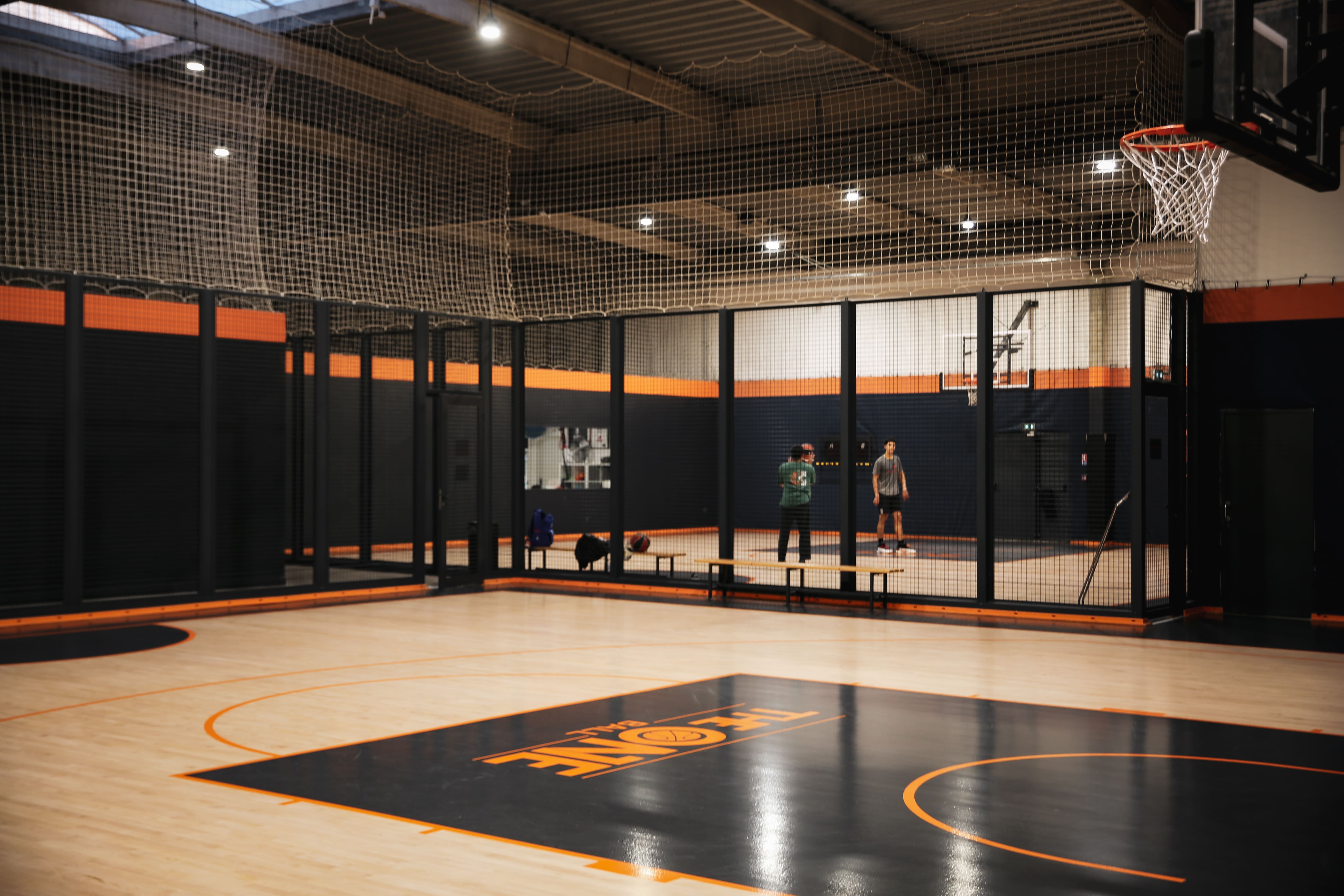 Deux personnes discutent sur un terrain de basketball intérieur avec un sol en bois clair et une zone centrale noire avec marquages orange.