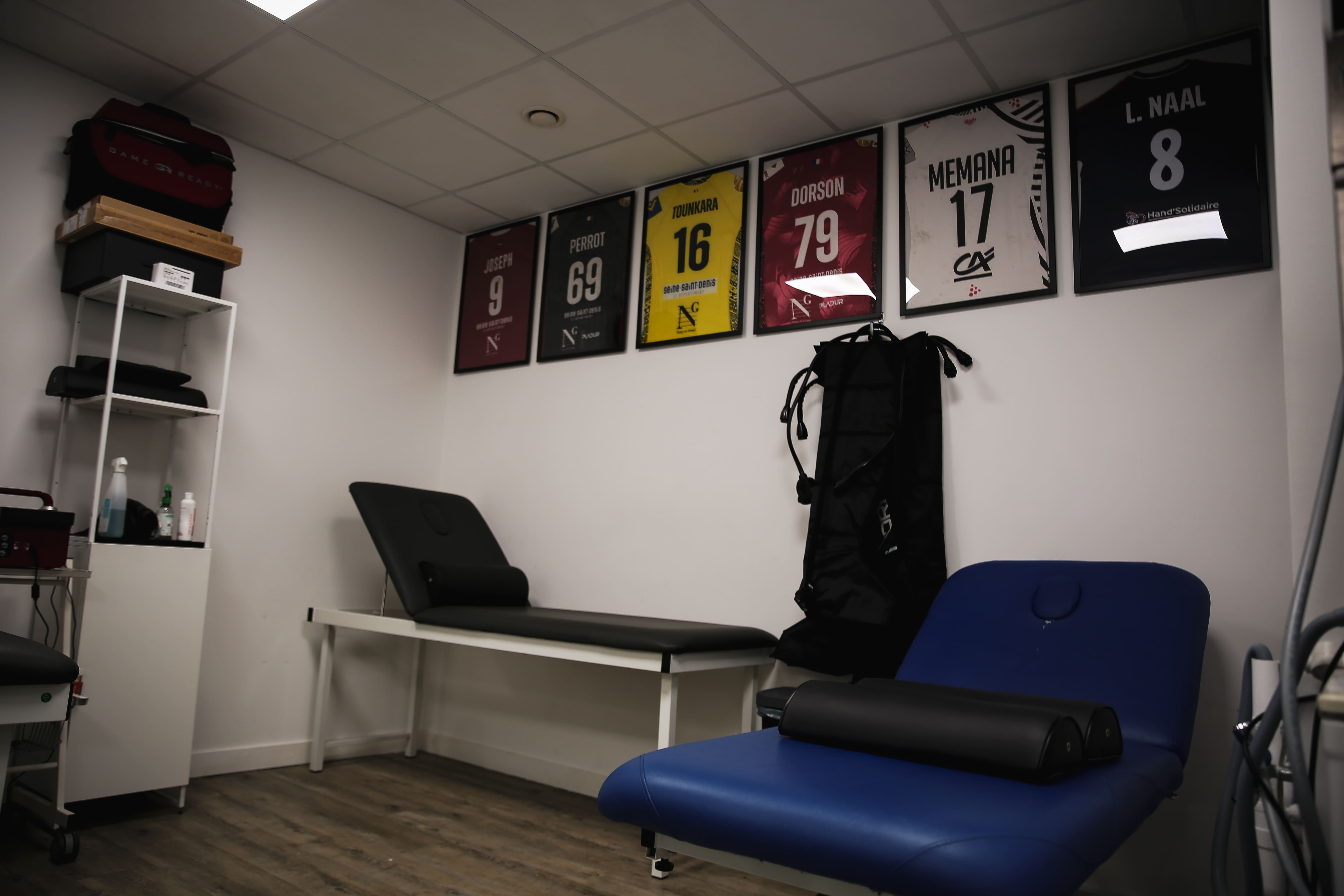 Salle de kinésithérapie avec deux tables de traitement, étagère blanche avec produits, et six maillots encadrés accrochés au mur.