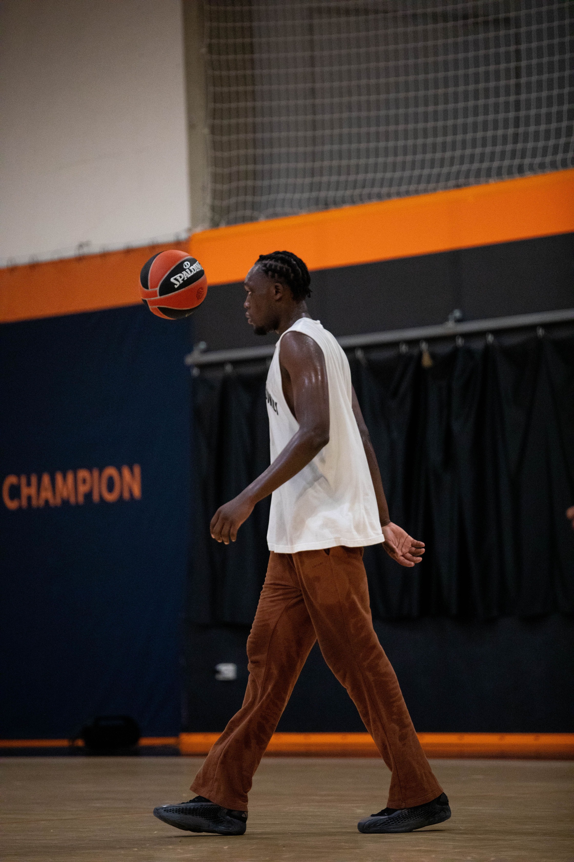 Un joueur de basketball portant un débardeur blanc et un pantalon marron marche sur un terrain intérieur avec un ballon Spalding en suspension devant lui.