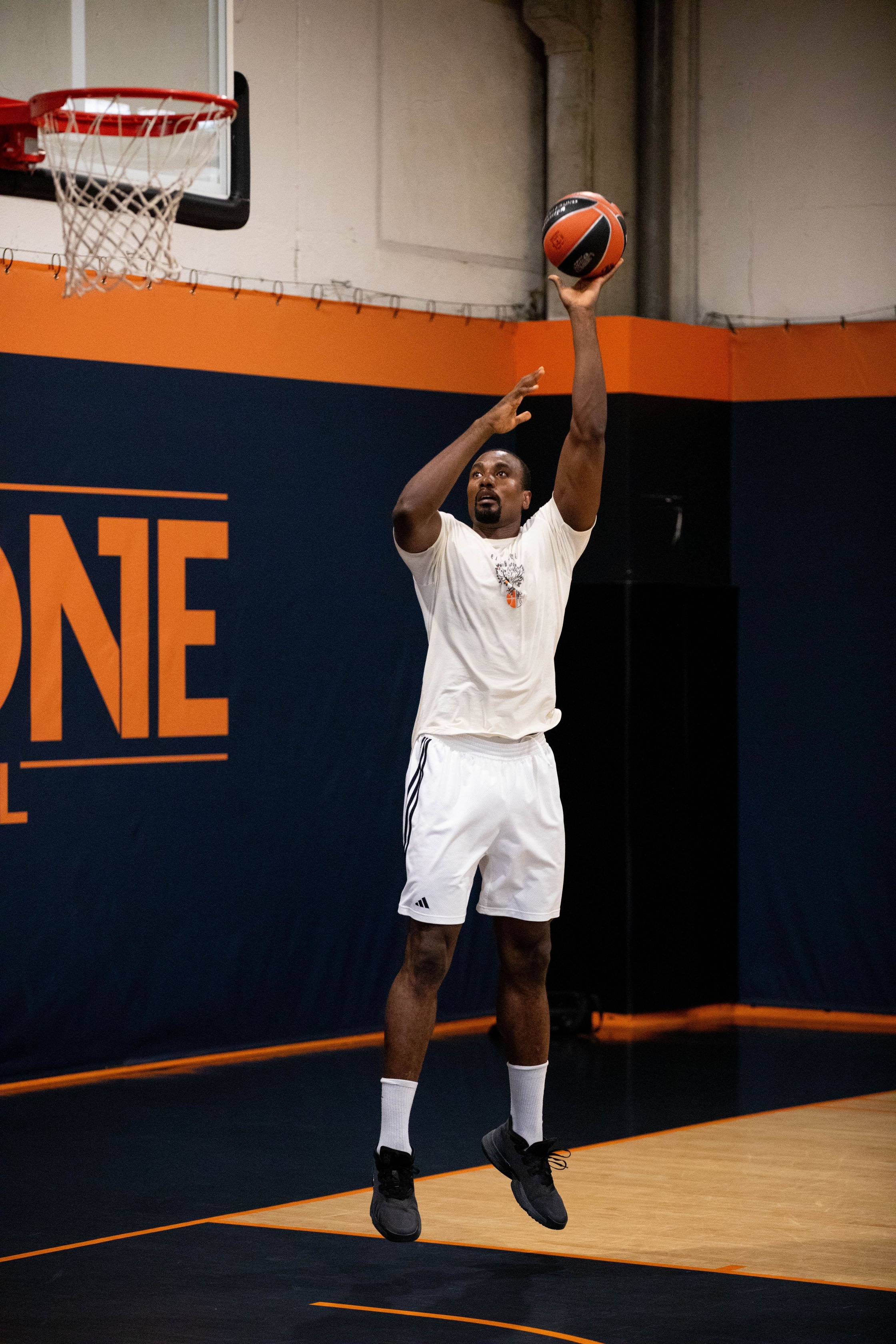 Homme en tenue blanche sautant pour tirer au basket-ball sur un terrain intérieur.