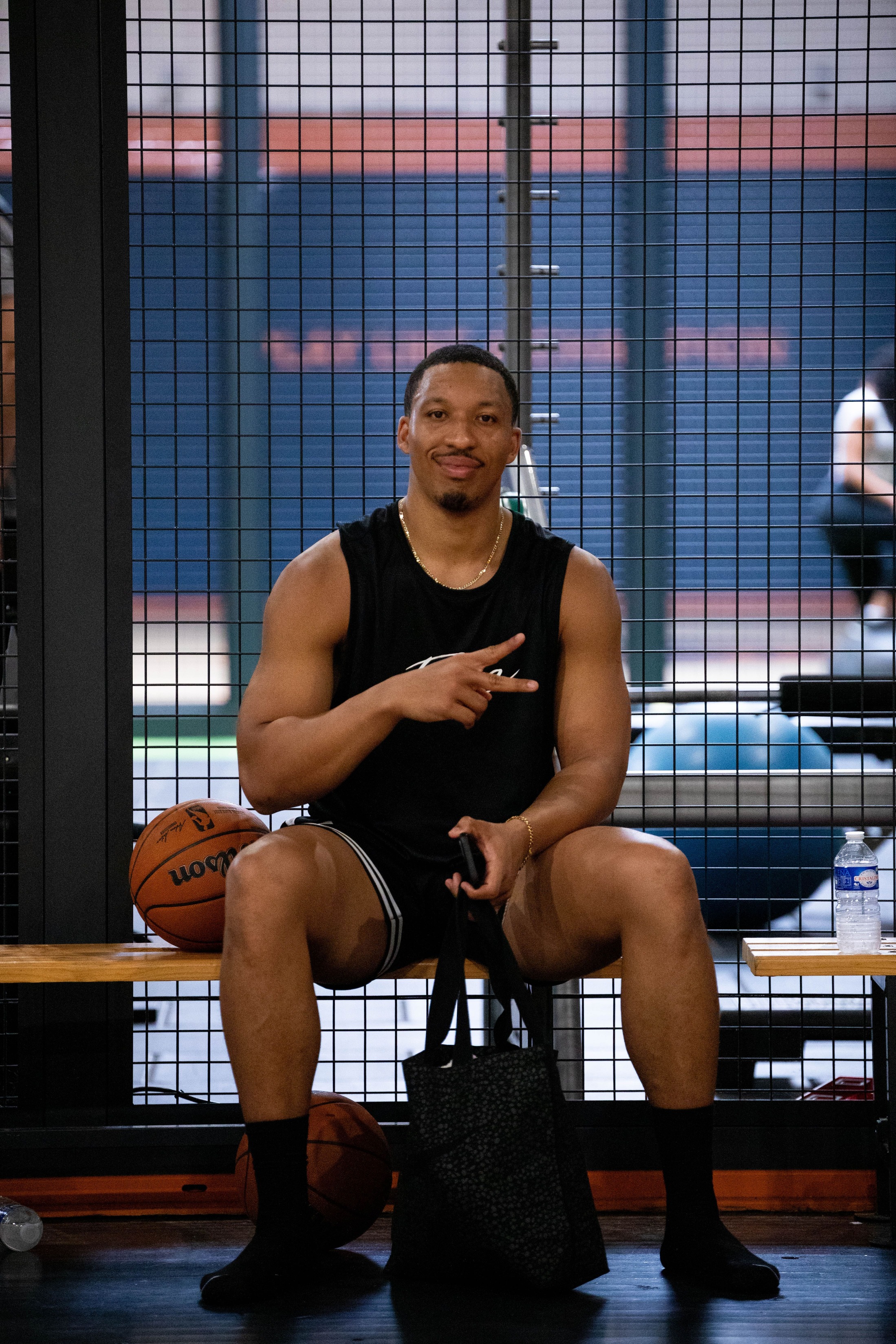 Homme assis sur un banc de gym, vêtu d'un débardeur noir et short, tenant un sac noir, avec deux ballons de basketball à proximité.