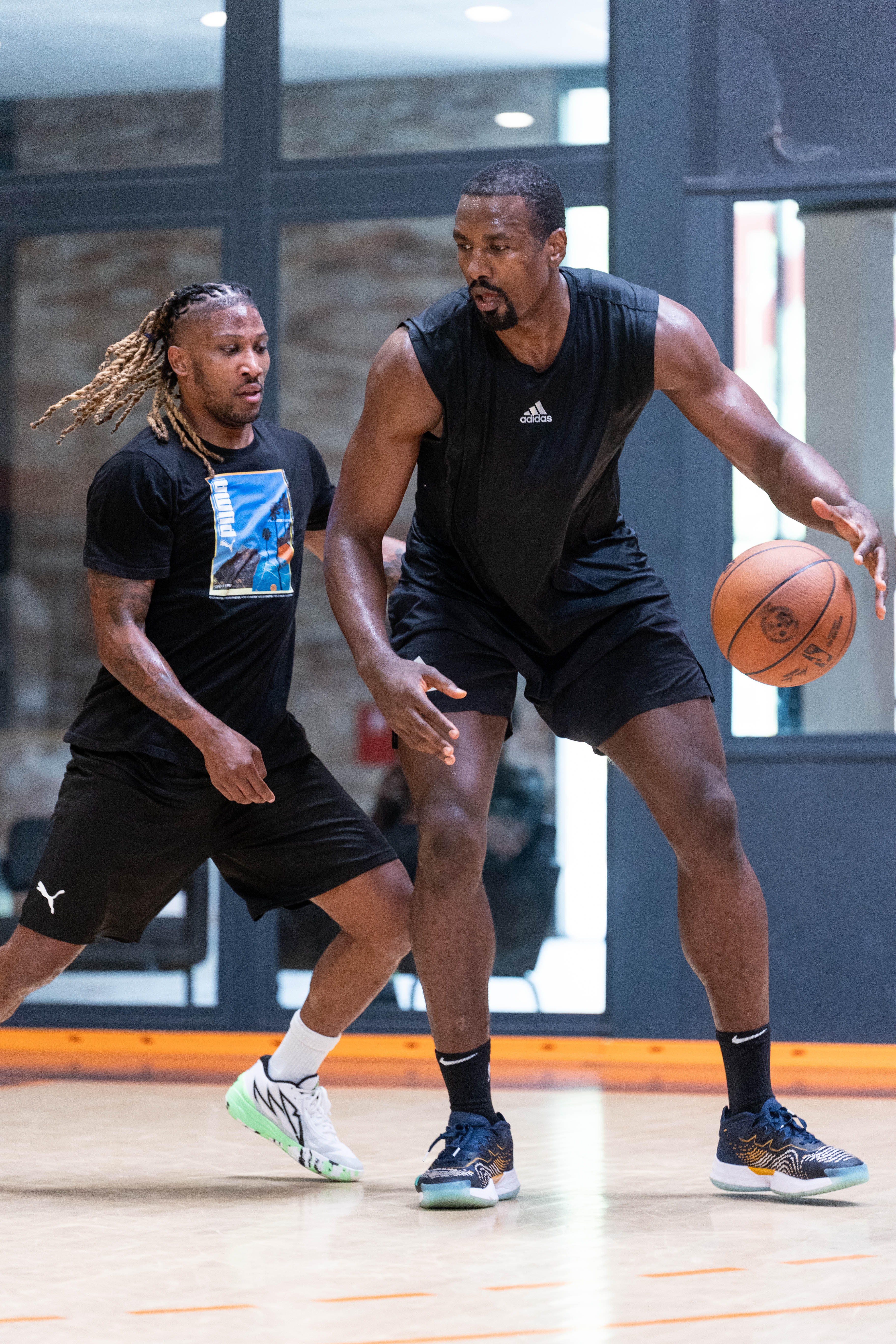 Deux hommes jouent au basketball en intérieur, l'un dribble le ballon tandis que l'autre défend de près.