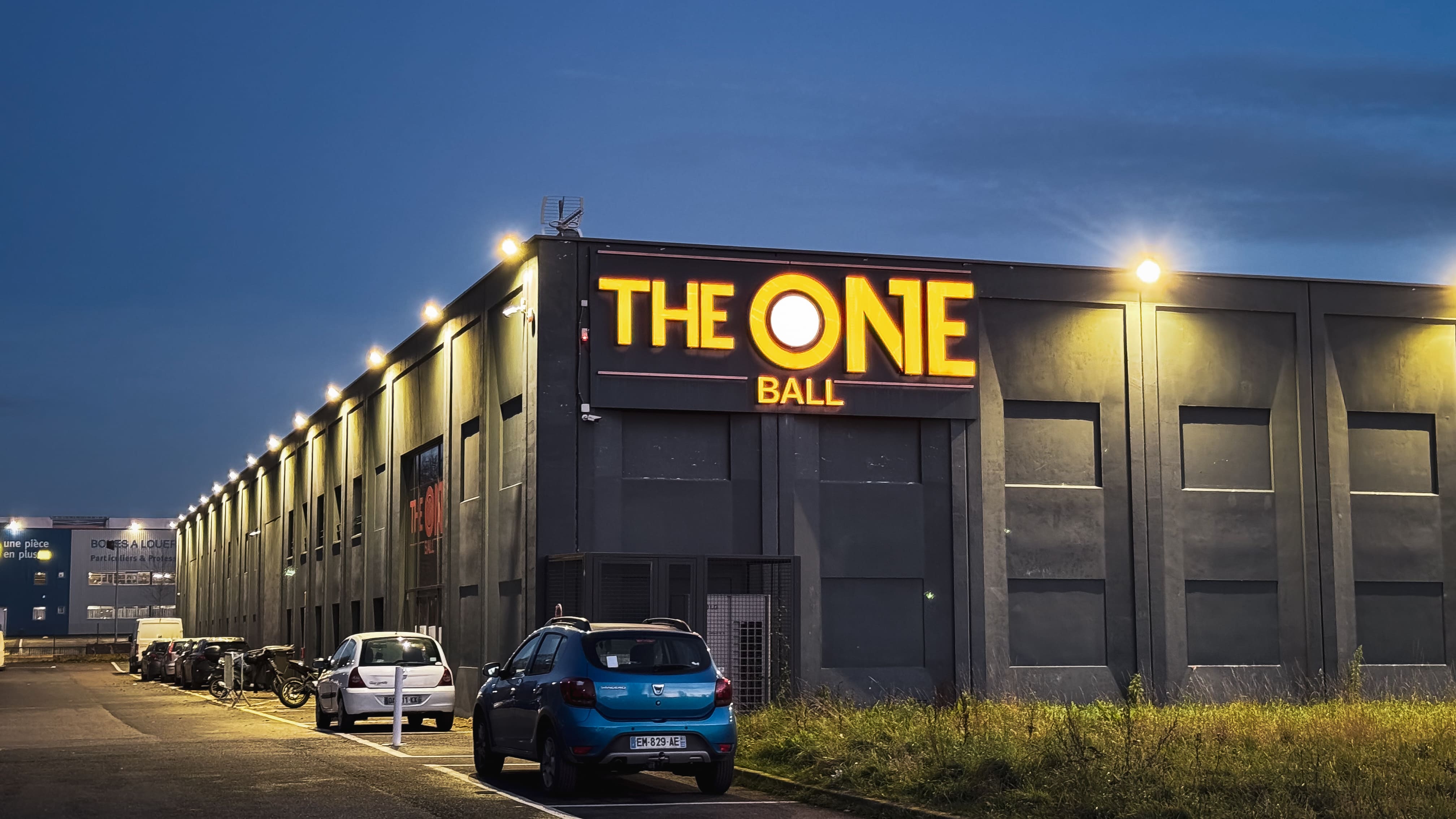 Façade d'un bâtiment industriel éclairé au crépuscule avec un panneau lumineux affichant le texte « THE ONE BALL » et plusieurs voitures stationnées devant.