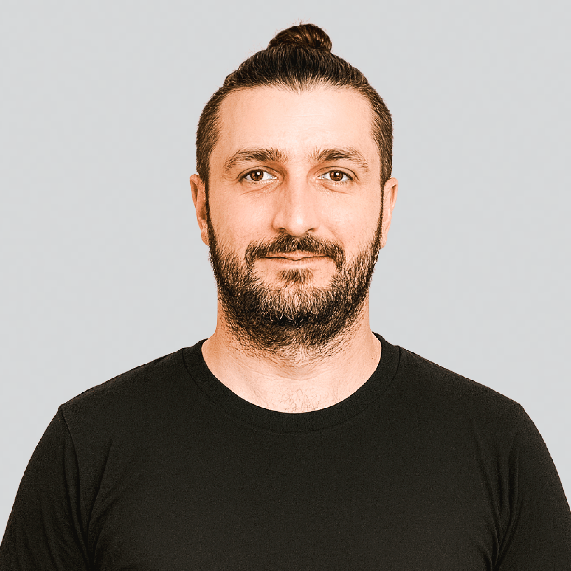 Man with a beard and mustache wearing a black t-shirt and a top knot hairstyle, against a light gray background.