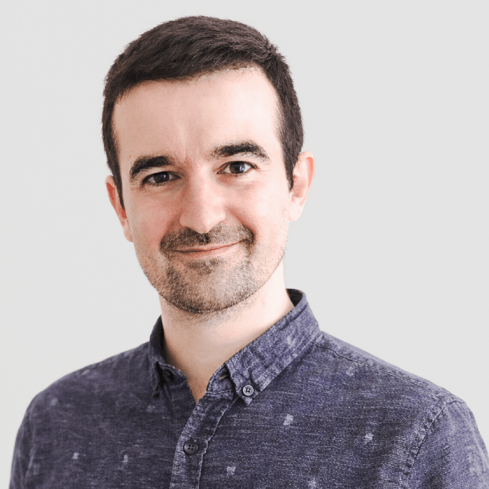 Smiling man with short dark hair and light facial hair wearing a blue patterned button-up shirt against a light background.