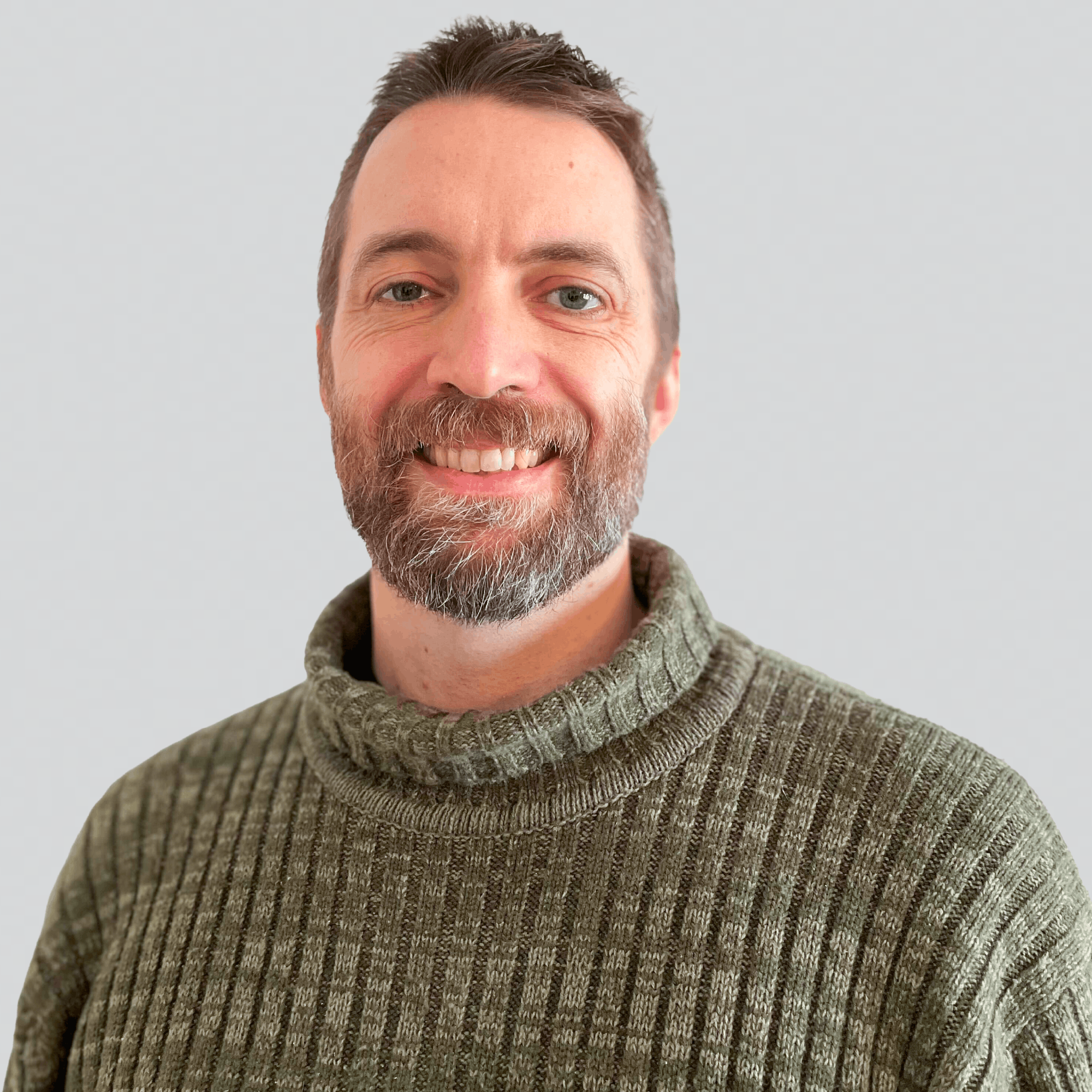 Smiling man with short brown hair and a beard wearing a green knit sweater against a light gray background.