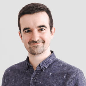 Smiling man with short dark hair and light facial hair wearing a blue patterned button-up shirt against a light background.