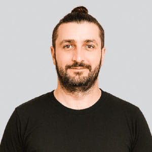 Man with a beard and mustache wearing a black t-shirt and a top knot hairstyle, against a light gray background.
