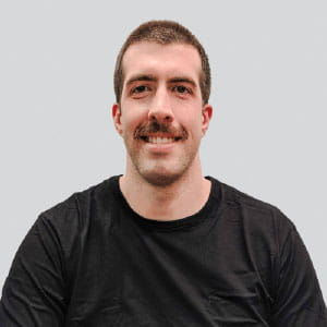 Smiling man with short brown hair and a mustache wearing a black t-shirt against a light gray background.