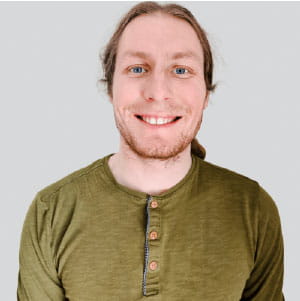 Smiling man with blue eyes and light brown hair wearing an olive green Henley shirt against a plain light gray background.
