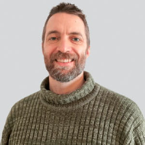 Smiling man with short brown hair and a beard wearing a green knit sweater against a light gray background.