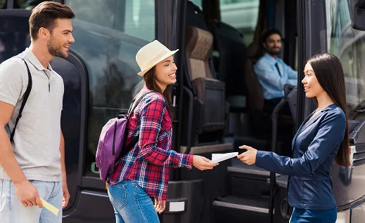 Young woman handing a ticket to a female bus attendant while a young man holds a ticket near a bus entrance with the driver seated inside.