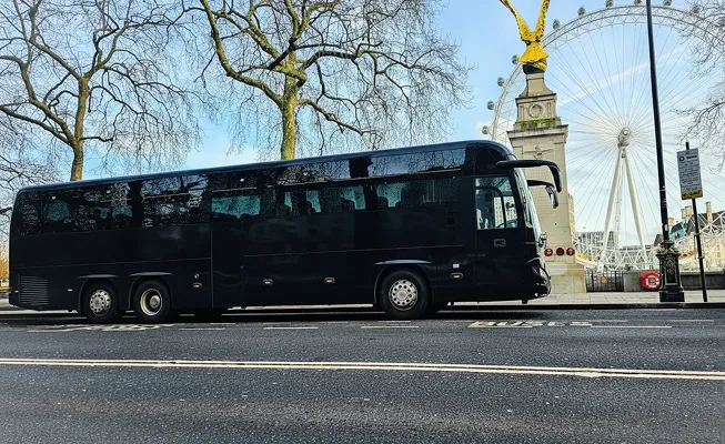 Black tour bus parked on a city street with trees, a stone pillar topped with a golden eagle, and the London Eye Ferris wheel in the background.