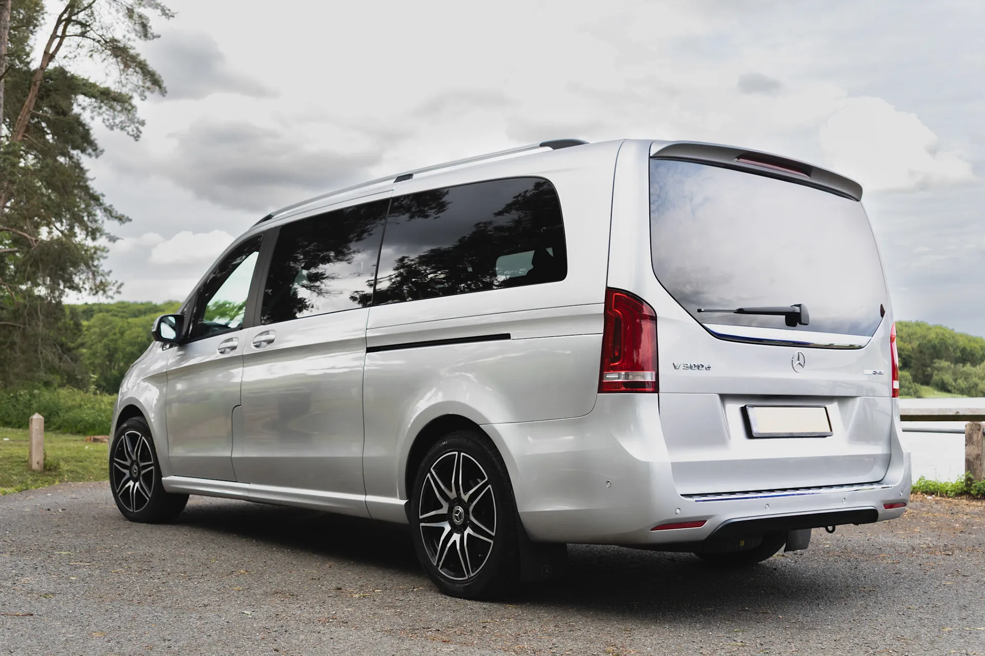 Silver Mercedes-Benz V-Class luxury van parked on a gravel road near forest and water.