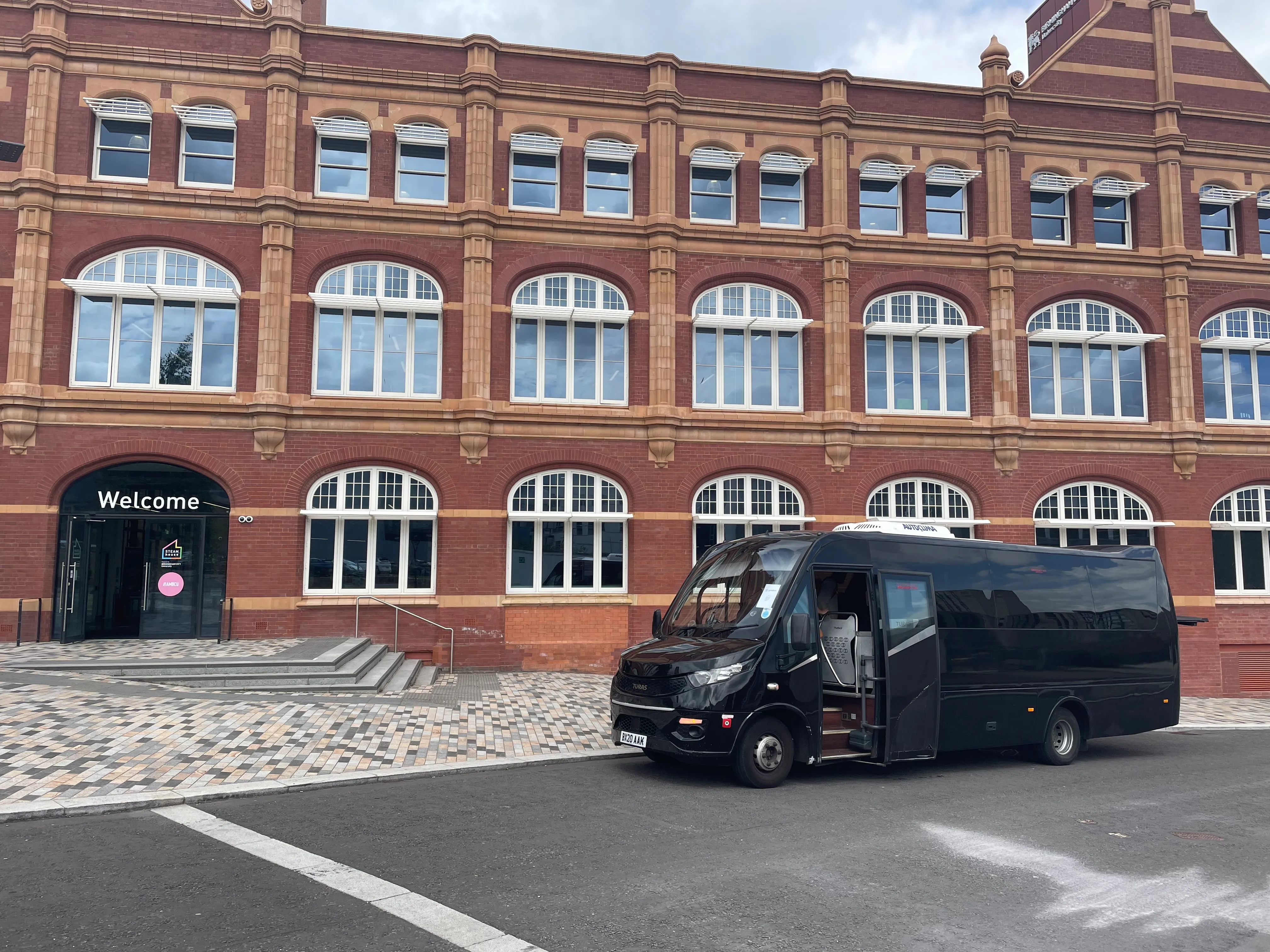 Black minibus parked in front of a red-brick building with arched windows and a 'Welcome' sign above the entrance.