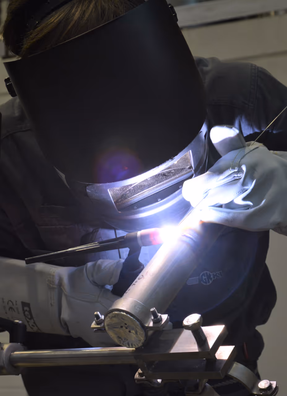 Person trägt Schweißhelm und Handschuhe und schweißt ein Metallrohr an einer Halterung.