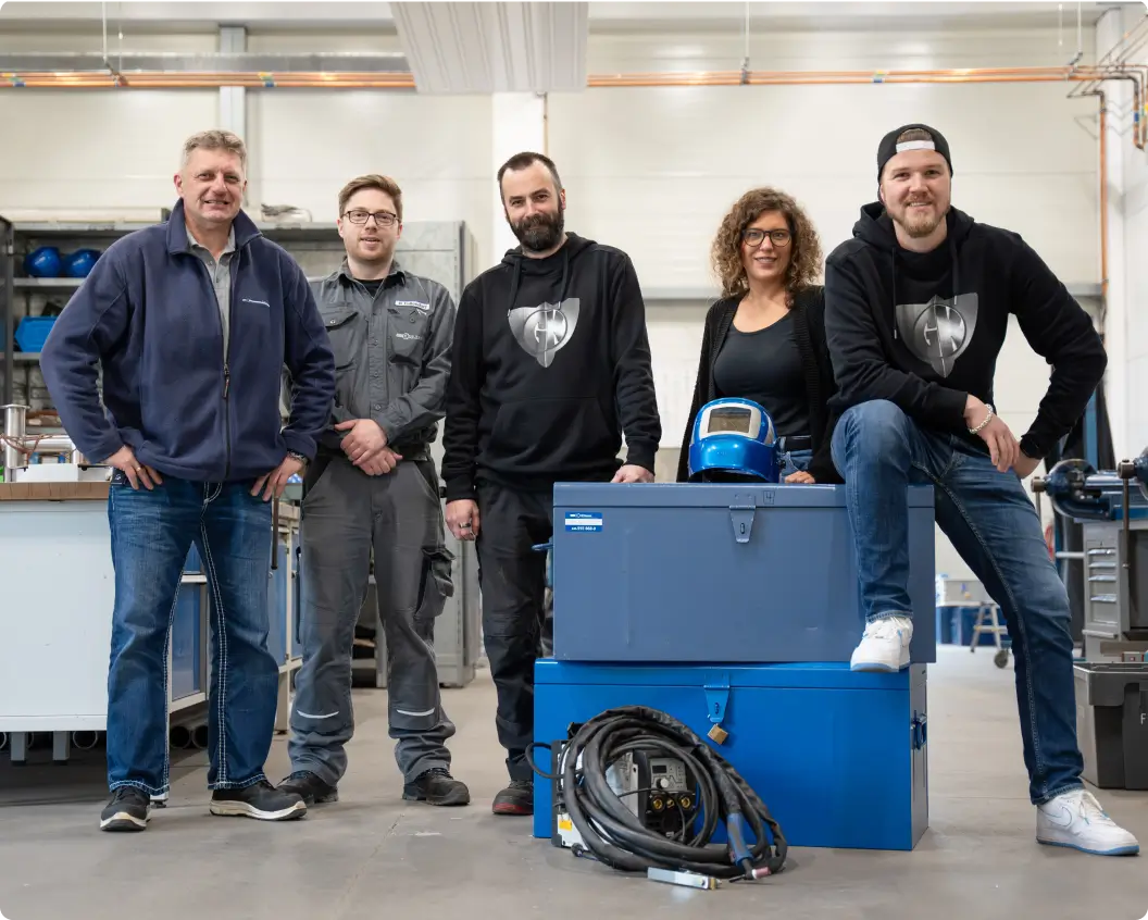 Fünf Mitarbeitende posieren in einer Werkstatt mit Ausrüstung zum Schweißen, darunter ein Schweißhelm und Kabel.