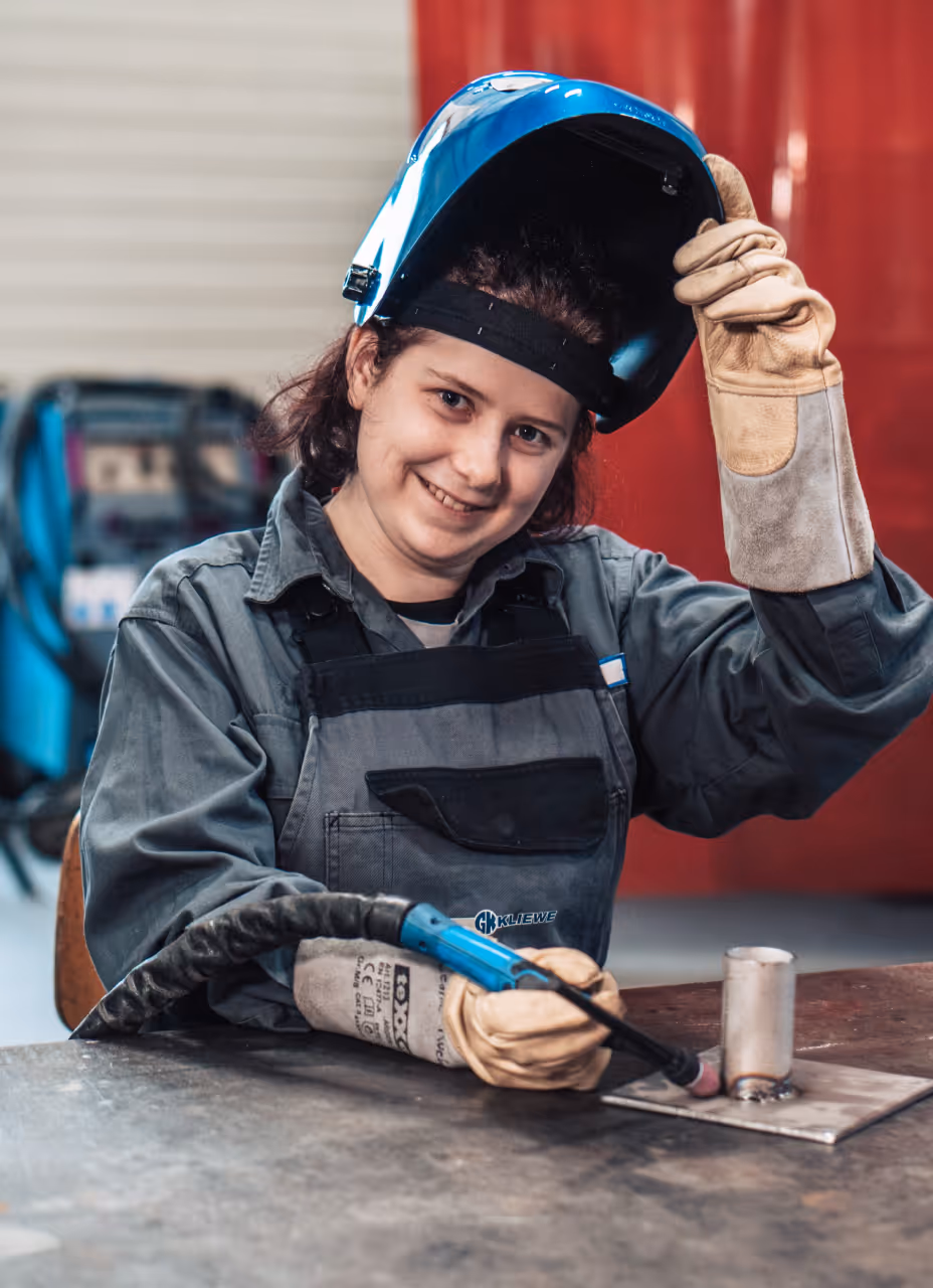 Person im Schweißeranzug hebt Schweißhelm und hält Schweißgerät über Metallplatte.