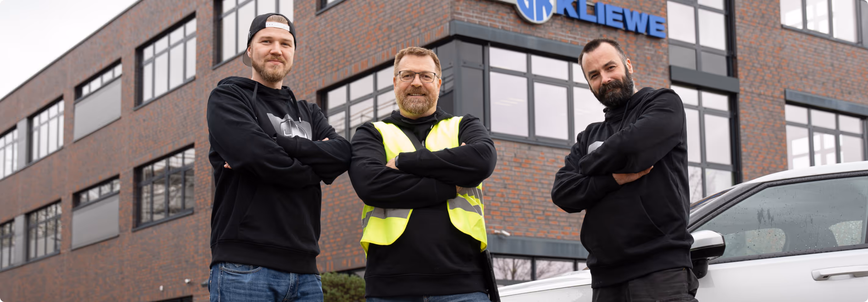 Drei Männer mit verschränkten Armen stehen vor einem modernen Bürogebäude und einem weißen Auto, einer trägt eine gelbe Sicherheitsweste.