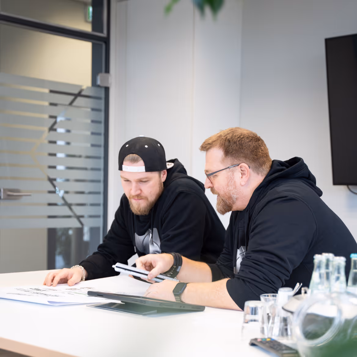 Zwei Männer in schwarzen Kapuzenpullovern arbeiten gemeinsam an Bauplänen an einem weißen Tisch in einem modernen Büro.