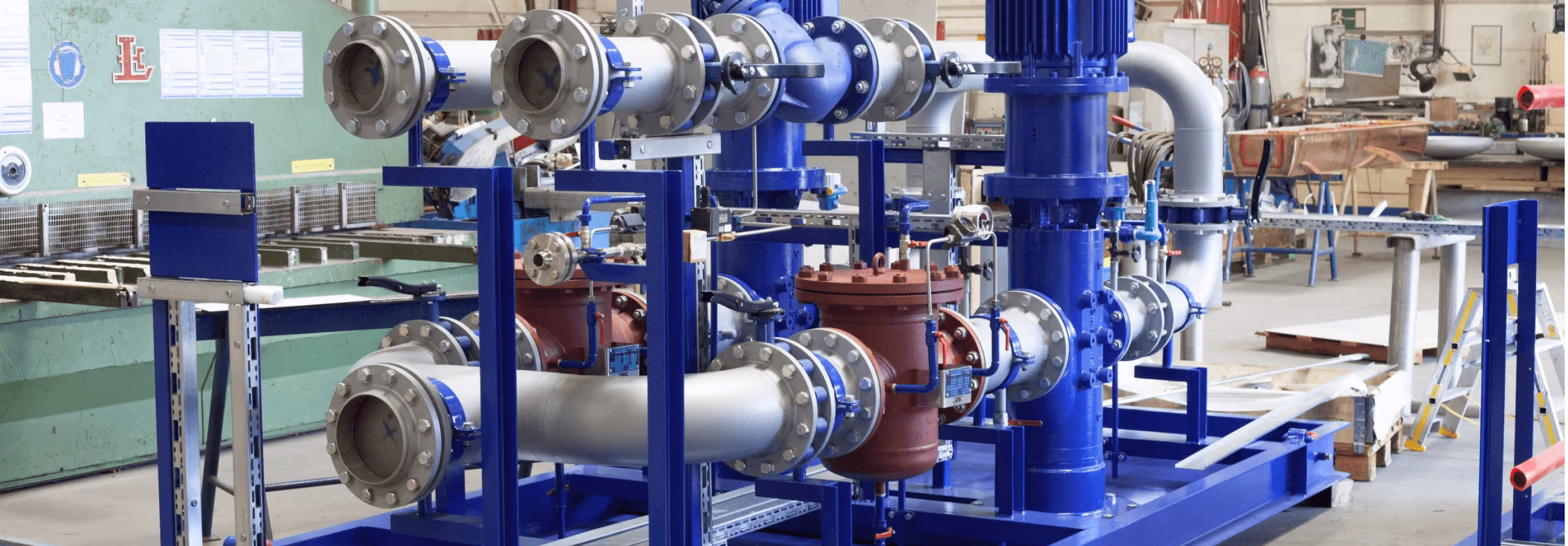 Industrielle Rohrleitung mit blauen und roten Metallventilen in einer Werkstatt mit Maschinen im Hintergrund.