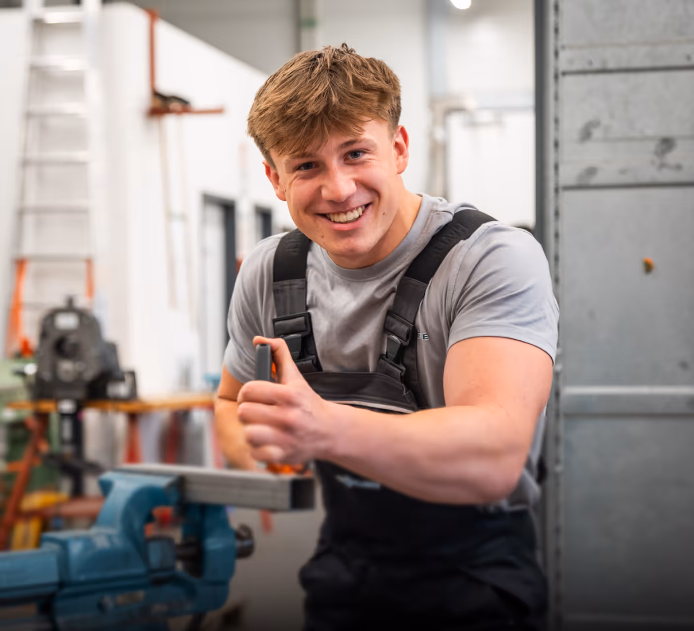 Junger Mann in Arbeitskleidung lächelt und hält eine Zange in einer Werkstatt.
