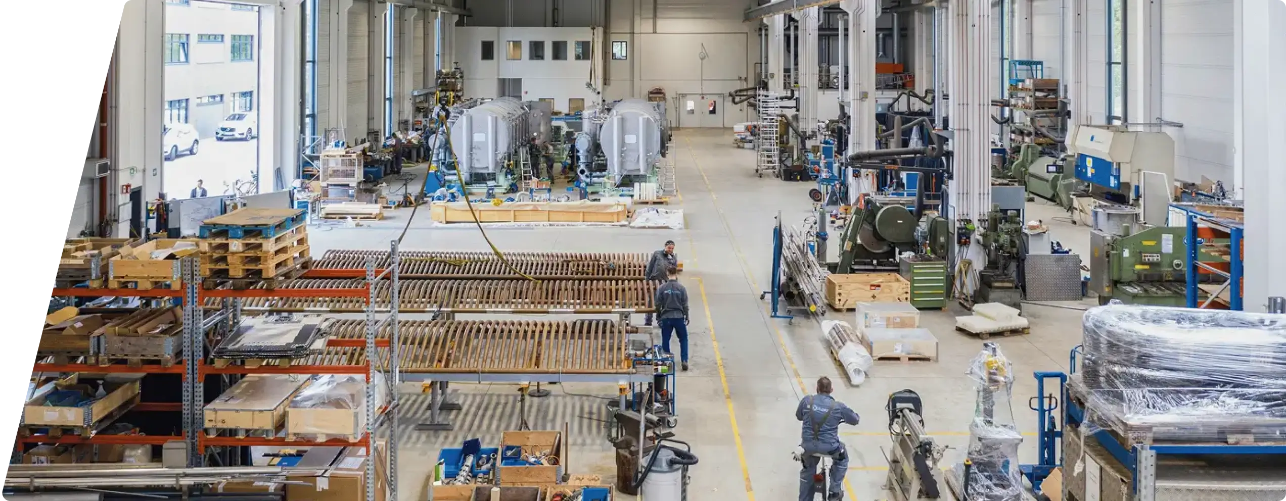 Große Industriehalle mit Maschinen, Lagerregalen und mehreren Arbeitern beim Montieren und Bewegen von Ausrüstung.