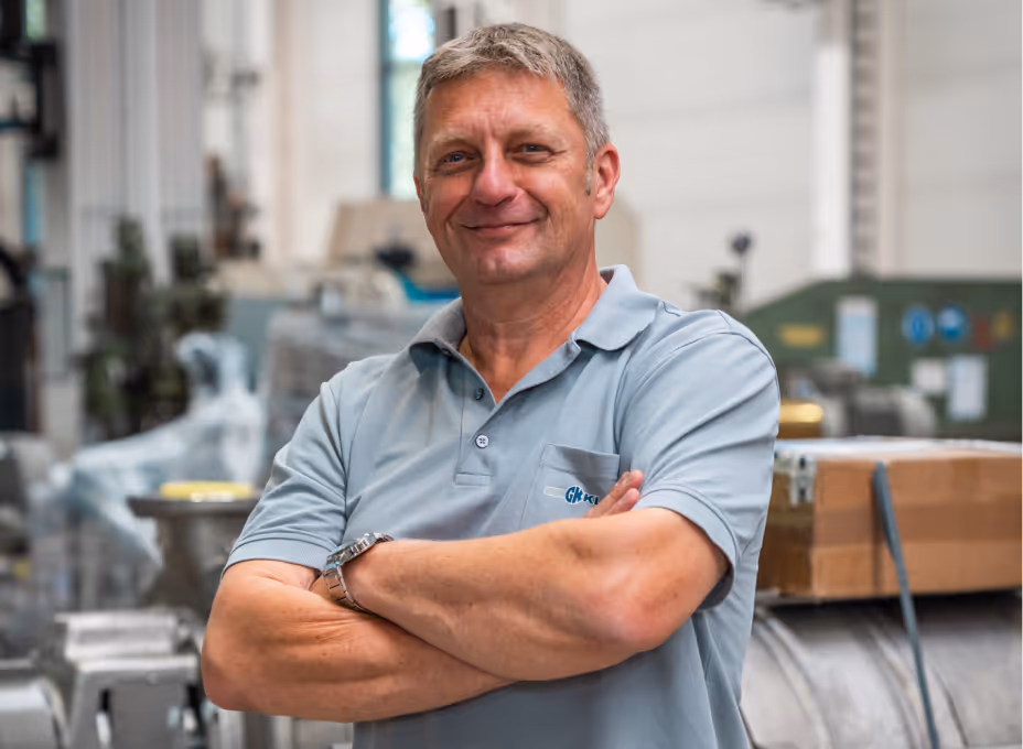 Mann in grauem Polohemd mit verschränkten Armen steht lächelnd in einer industriellen Werkstatt.