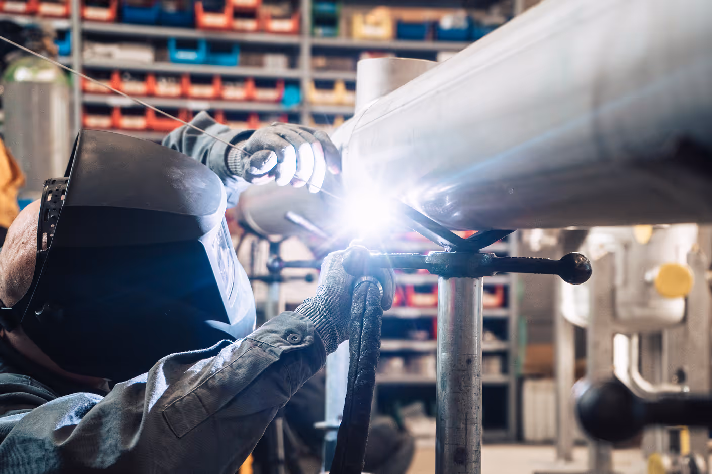 Schweißer mit Schutzhelm und Handschuhen schweißt ein großes Metallrohr in einer Werkstatt mit Werkzeugregalen im Hintergrund.