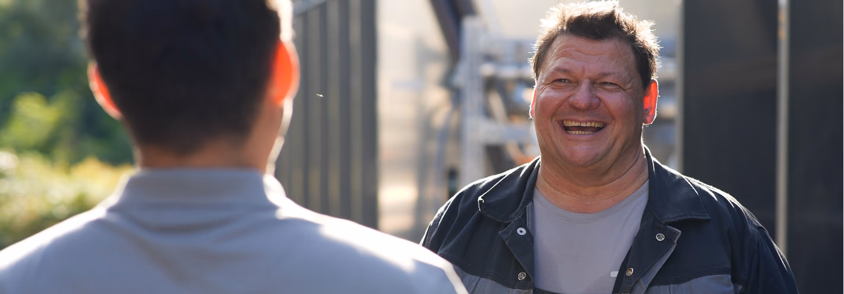 Zwei Männer unterhalten sich draußen, einer lächelt freundlich in die Kamera.