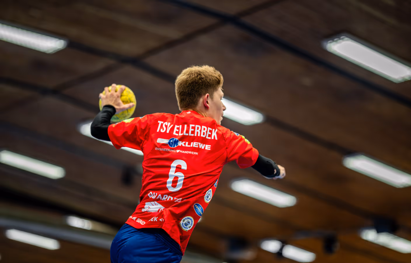 Handballspieler mit rotem TSV Ellerbek Trikot wirft einen gelben Ball in der Halle.