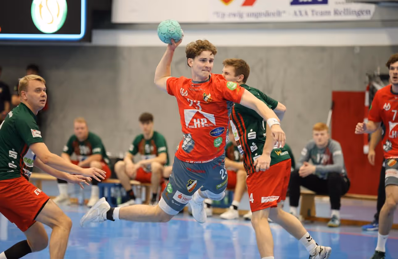 Handballspieler in rotem Trikot springt zum Wurf, umgeben von Gegnern in grünen Trikots auf der Halle.