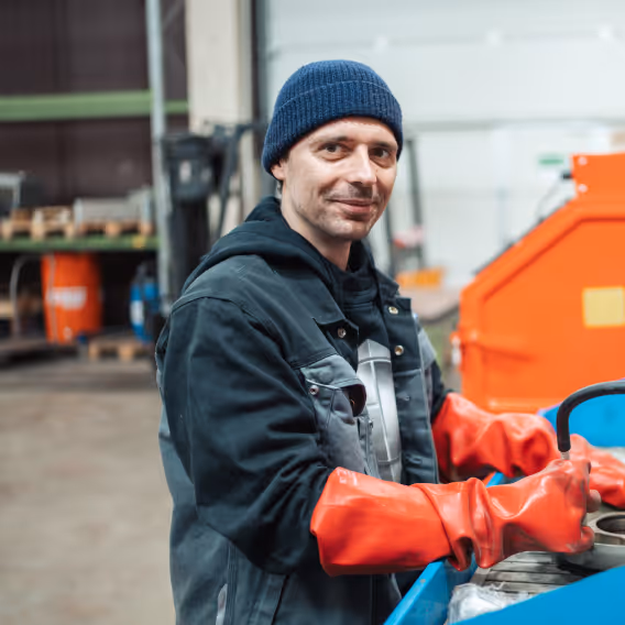 Ein Mann in Arbeitskleidung und blauer Mütze arbeitet mit roten Handschuhen an einer Maschine in einer Industriehalle.