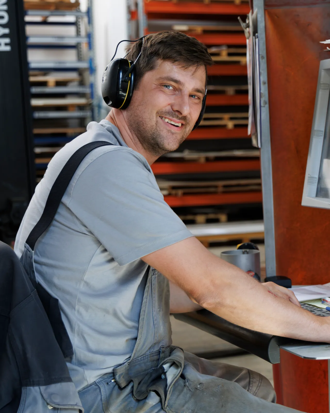 Lächelnder Mann in Arbeitskleidung mit Gehörschutz sitzt an einem Schreibtisch in einer Werkstatt.