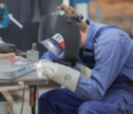 Person in blauer Arbeitskleidung und Schutzhelm bedient eine Schweißmaschine in einer Werkstatt.